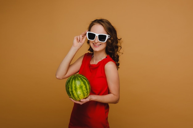 Een mooi meisje in glazen en een rode jurk heeft een watermeloen in haar handen