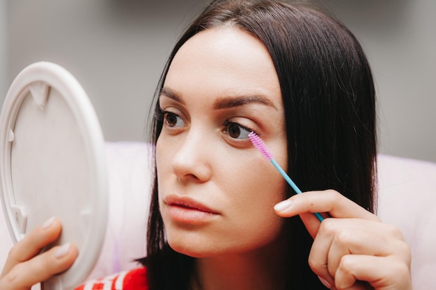 Een mooi meisje kijkt in de spiegel en brengt make-up aan, de vrouw heeft haar wimpers vergroot en kijkt in de spiegel