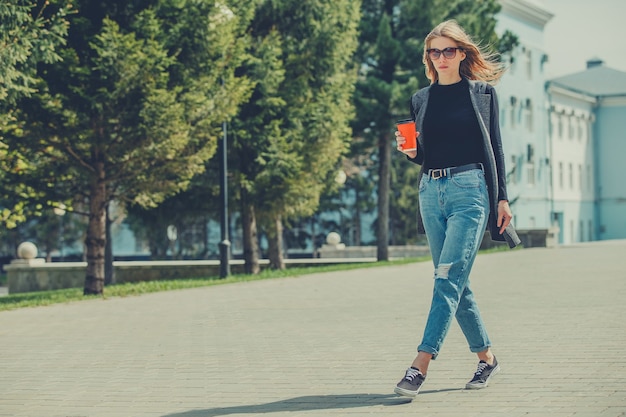 Een mooi meisje loopt onderweg over straat met een kop koffie.