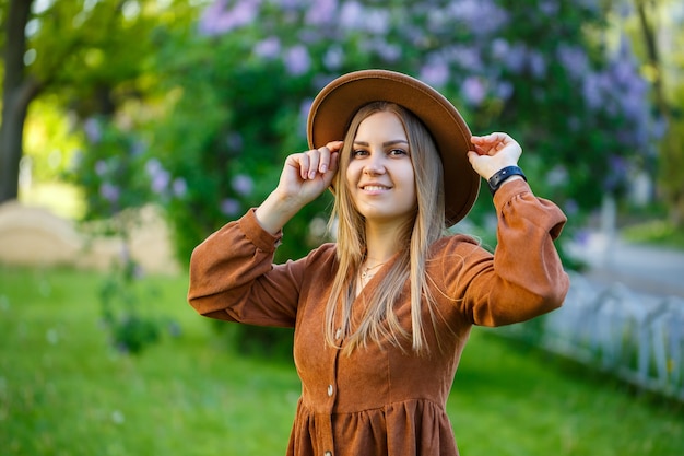 Een mooi meisje met blond haar houdt een hoed in haar handen en staat in de buurt van een lila struik. Jonge vrouw in de tuin met bloeiende bomen