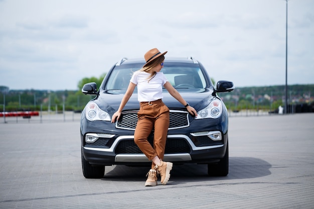 Een mooi meisje met een bril en een bruine hoed staat in de buurt van een zwarte auto. jonge vrouw met auto op de parkeerplaats