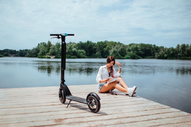 Een mooi meisje met een elektrische scooter leest een boek op een houten brug bij de rivier. eco vervoer en gezondheidszorg concept.