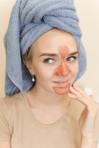 Een mooi meisje met een handdoek op haar hoofd maakt een multimasker op de t-zone van oranje kleur met een scrub.