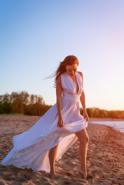 Een mooi meisje met een Kaukasisch uiterlijk staat alleen in een lange witte jurk aan de kust in de w...