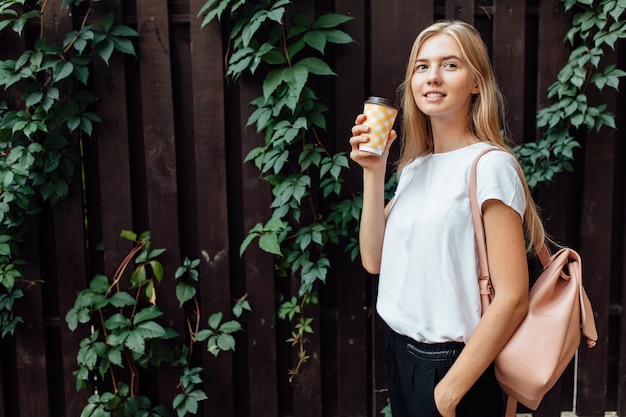 Een mooi meisje met een kopje koffie, gekleed in een wit t-shirt, staat in de open lucht, aan de houten loofmuur, drinkt een drankje