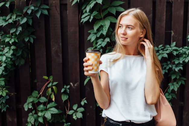 Een mooi meisje met een kopje koffie, gekleed in een wit t-shirt, staat in de open lucht, aan de houten loofmuur, drinkt een drankje