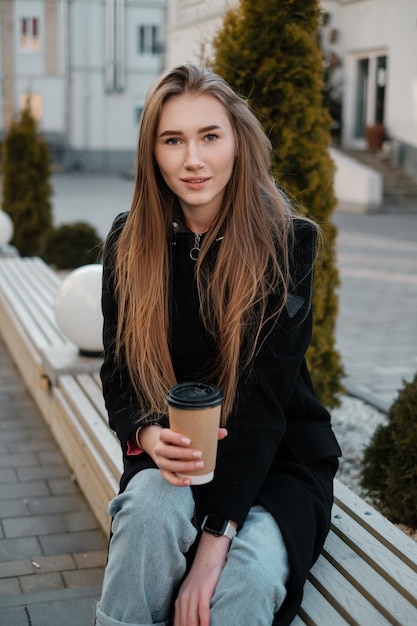 Een mooi meisje met lang haar met koffie zittend op een bankje en wegkijkend Casual straat vrouwelijk portret