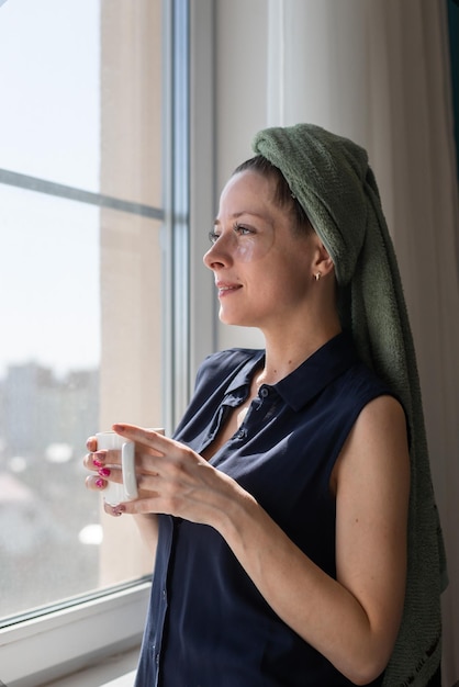 Een mooi meisje na een douche in een handdoek op haar hoofd staat thuis kijkt uit het raam en houdt een mok warme drank vast De tijd is niet alleen Persoonlijke verzorging