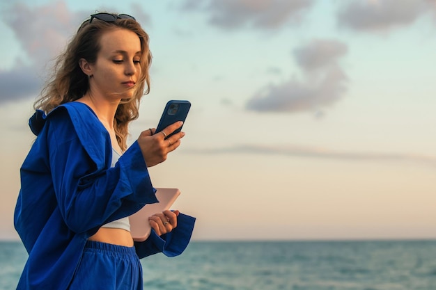 een mooi meisje staat aan de kust van de avondzee, in de handen van een tablet en een telefoon