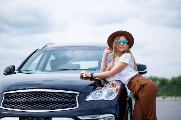 Een mooi meisje van Europees uiterlijk met een bril en een bruine hoed staat in de buurt van een zwarte auto. Fotoshoot bij de auto.