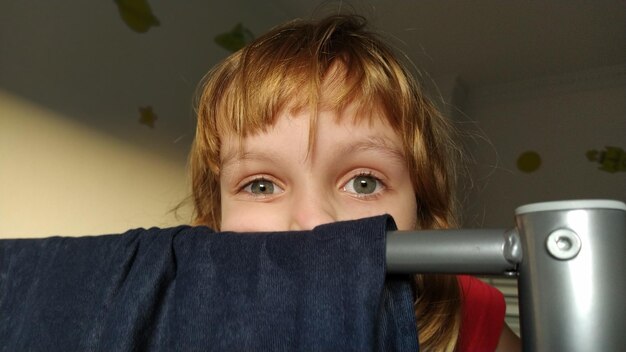 Een mooi meisje verstopt zich achter de rug van een stapelbed waaraan donkerblauwe fluwelen stof hangt het kind heeft grijze ogen en blond haar de bovenste helft van het gezicht is zichtbaar verstoppertje