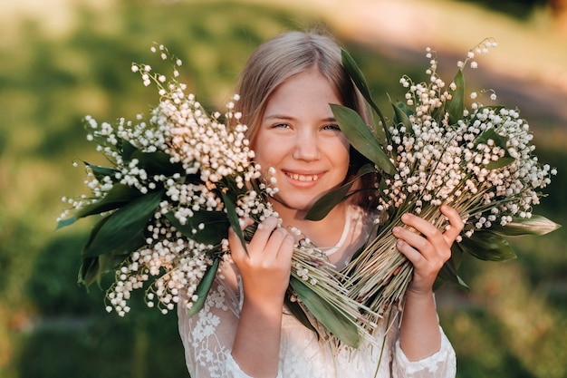 Een mooi negenjarig blond meisje met lang haar in een lange witte jurk, met een boeket van lelietje-van-dalenbloemen, wandelend in de natuur in het park.summer, zonsondergang.