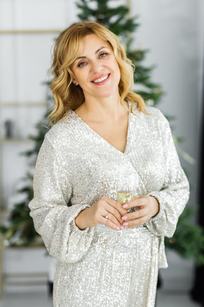 Een mooi rijp blond meisje in een gouden jurk poseert in de buurt van de kerstboom met een glas champagne in haar handen Vrouw lacht en kijkt naar de camera