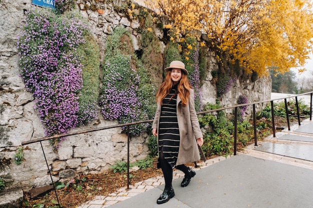 Een mooi romantisch meisje in een jas en hoed loopt in Annecy. Frankrijk. Meisje in een hoed in Frankrijk op de achtergrond van bloeiende planten