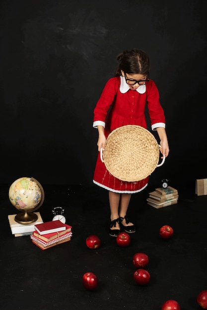 Een mooi schoolmeisje in een rode jurk met een boeken appels en globe op een zwarte achtergrond