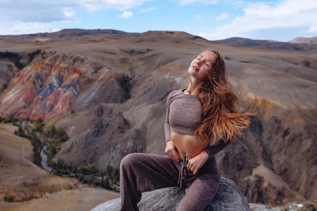 Een mooi slank meisje in een trainingspak zit op een rots met haar ogen gesloten tegen de achtergrond van de rotsachtige bergen