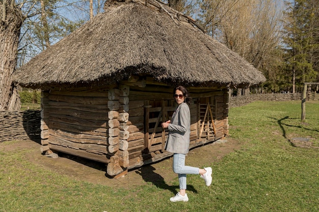 Een mooi stijlvol gekleed meisje tegen de achtergrond van een oud landelijk huis onder een rieten dak.