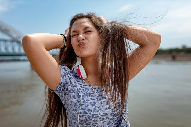 Een mooi vrolijk meisje dat pruilt, een gezicht trekt en plezier heeft met je haar in de open lucht en geniet op de stadsoever van de rivier op een mooie zomerdag.