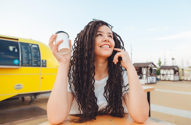 Een mooi vrolijk meisje met dreadlocks zit op een food court en drinkt koffie fastfood café op straat een vrouw drinkt koffie in een café herfst