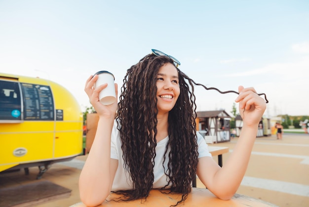 Een mooi vrolijk meisje met dreadlocks zit op een food court en drinkt koffie fastfood café op straat een vrouw drinkt koffie in een café herfst