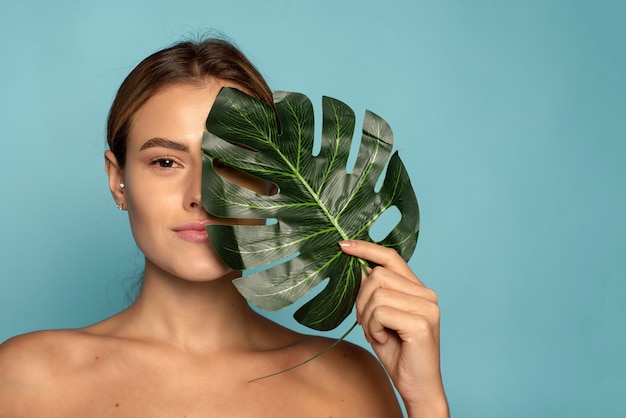 Een mooi vrouwelijk model bedekt een deel van haar gezicht met een tropisch palmblad. huidverzorging, hydratatie. cosmetica met natuurlijke ingrediënten.