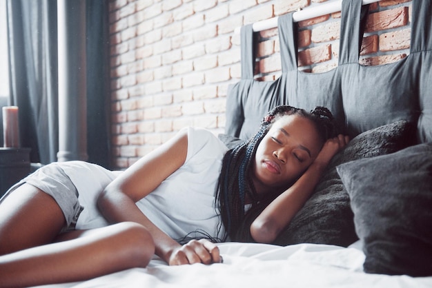 Een mooie Afrikaanse vrouw ligt en slaapt in bed in een witte slaapkamer Goedemorgen vakantie thuis het meisje draagt een T-shirt pigtails op haar hoofd