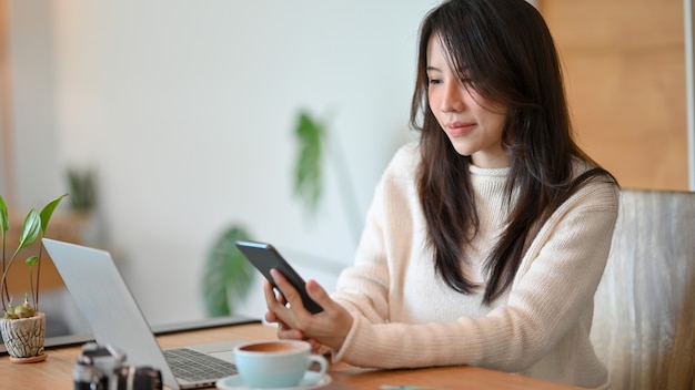 Een mooie Aziatische jongedame zit in de co-workingruimte van het café en leest online zakelijk nieuws op haar smartphone.