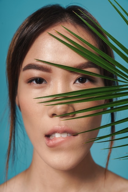 een mooie Aziatische serieuze vrouw poseren naakt geïsoleerd over blauwe muur muur bedrijf plant blad bijten haar lip.