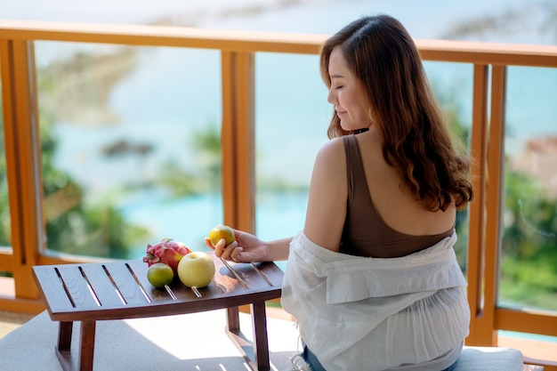 Een mooie aziatische vrouw die fruit vasthoudt terwijl ze geniet van het zitten op het balkon met de achtergrond van het uitzicht op zee