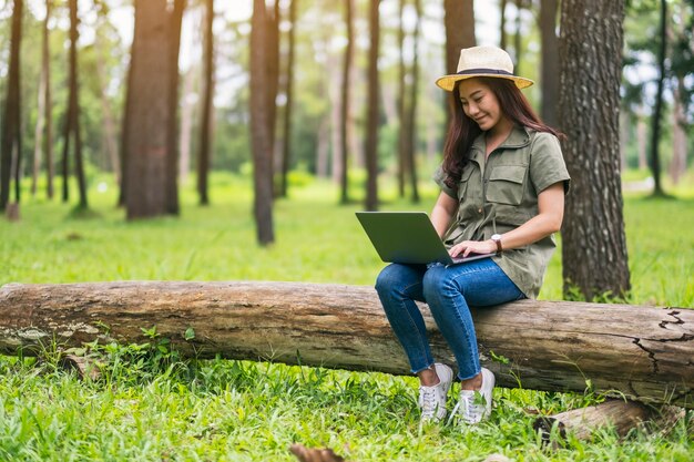 Een mooie aziatische vrouw die het toetsenbord van een laptop gebruikt en typt terwijl ze op een boomstam in het park zit