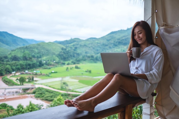 Een mooie aziatische vrouw die op een laptop werkt en typt terwijl ze op een balkon zit met bergen en groene natuurachtergrond