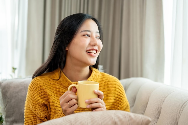 Een mooie Aziatische vrouw houdt een koffiebeker vast en zit op een bank te glimlachen en te dagdromen.