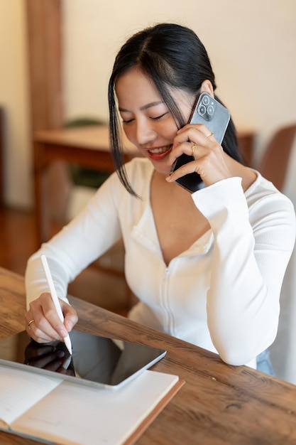 Een mooie Aziatische vrouw is aan de telefoon terwijl ze haar tablet gebruikt terwijl ze op afstand werkt in een café