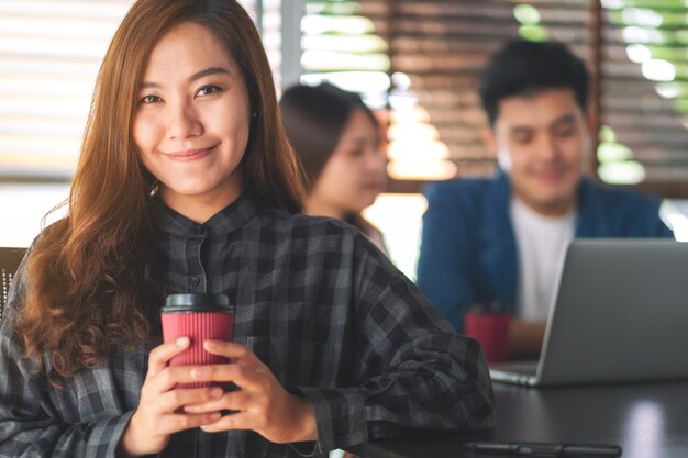 Een mooie aziatische vrouw met een papieren koffiekopje met collega's die op de achtergrond aan een laptop werken