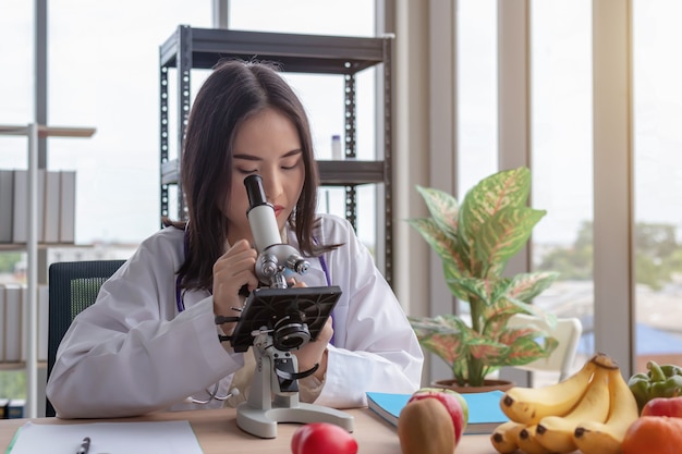 Een mooie Aziatische vrouwelijke arts bekijkt een microscoop op een modern bureau met een groot glasvensterachtergrond.