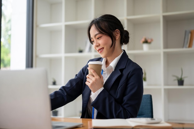 Een mooie Aziatische zakenvrouw drinkt koffie terwijl ze aan haar taken werkt op haar laptop.