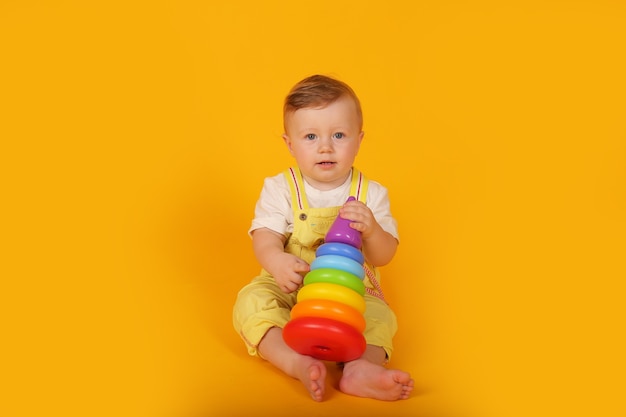 Een mooie, blauwogige kleine jongen in een geel pak speelt een veelkleurige speelgoedpiramide