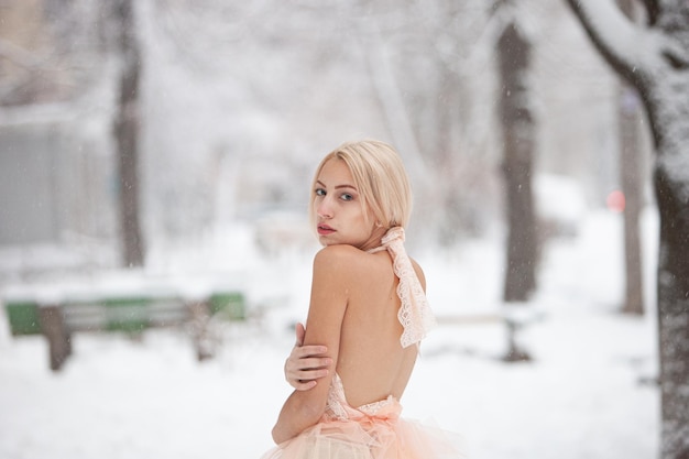 Een mooie blonde in een chique roze jurk tegen de achtergrond van een besneeuwd park