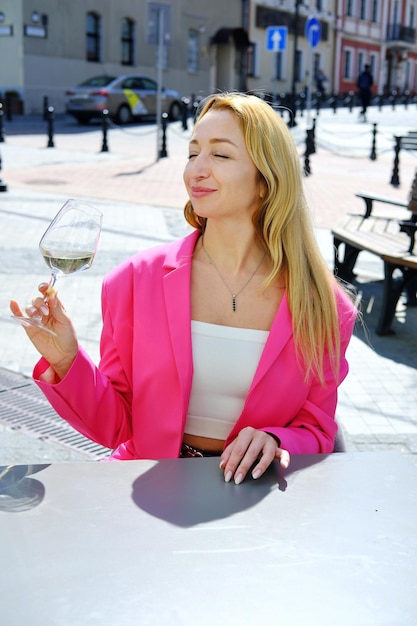 Een mooie blonde volwassen vrouw in een fel fuchsiakleurig jasje zit aan een tafel in een café, drinkt witte wijn en geniet van het weer