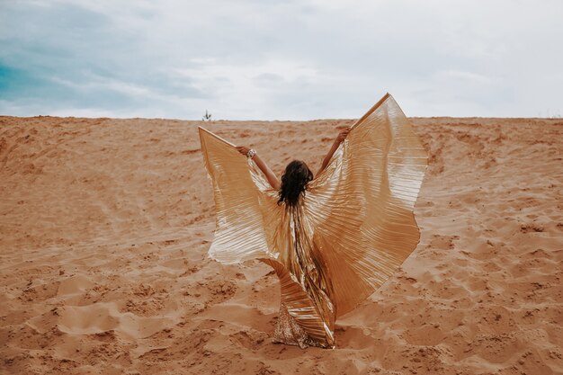 Een mooie blonde vrouw in een gouden met schitterende jurk met vleugels, kostuum danst een oosterse, oosterse dans in de woestijn op het zand. Mooie exotische vrouwendanser in een kostuum voor buikdansen.