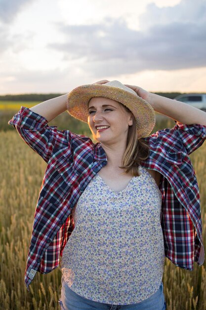 Een mooie boerenvrouw van middelbare leeftijd in een strohoed en een geruit hemd staat overdag in het zonlicht in een veld van gouden rijpende tarwe