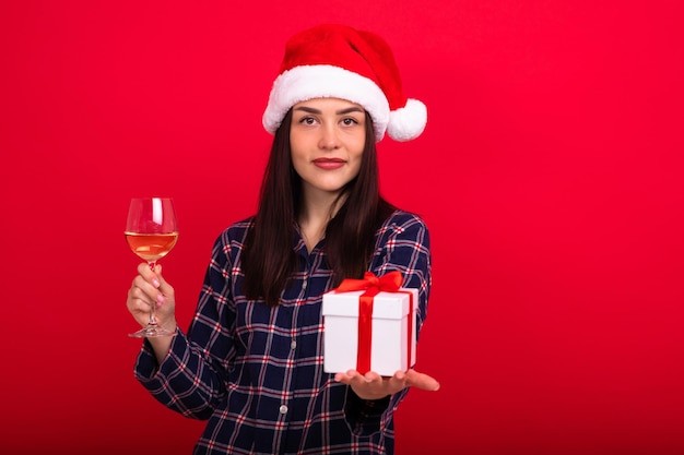 Een mooie brunette in pyjama in een kerstmanhoed houdt een glas wijn en een geschenkdoos op een rode achtergrond vrolijk kerstfeest