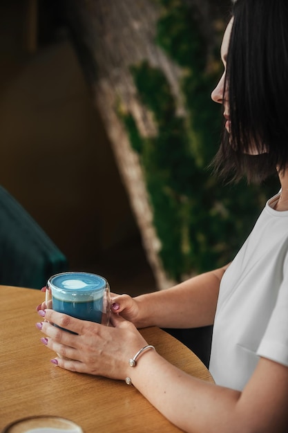 Een mooie brunette vrouw in een witte blouse zit in een café en drinkt blauwe Thaise thee