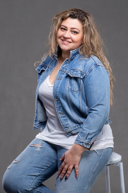 Een mooie dikke vrouw in een spijkerjasje kijkt naar de camera en glimlacht op een grijze achtergrond.
