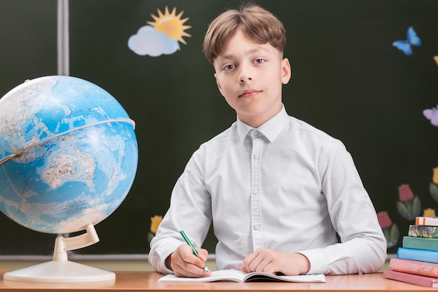 Een mooie gelukkige schooljongen zit aan een bureau op de achtergrond van een bord met stickers