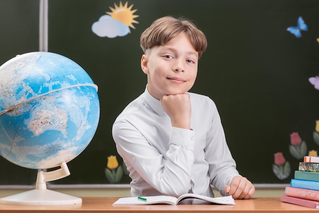 Een mooie gelukkige schooljongen zit aan een bureau op de achtergrond van een bord met stickers