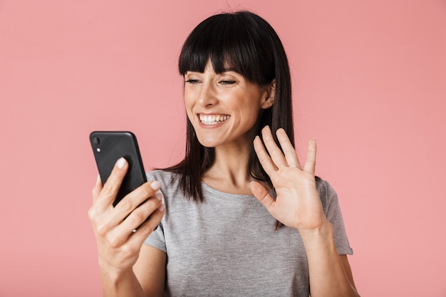 een mooie geweldige opgewonden gelukkige vrouw poseren geïsoleerd over licht roze muur muur met behulp van mobiele telefoon praten zwaaien.