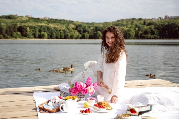 Een mooie glimlachende jonge vrouw met lang krullend haar gekleed in een witte jurk die een kopje thee serveert, gaat picknicken in een zonnige tijd