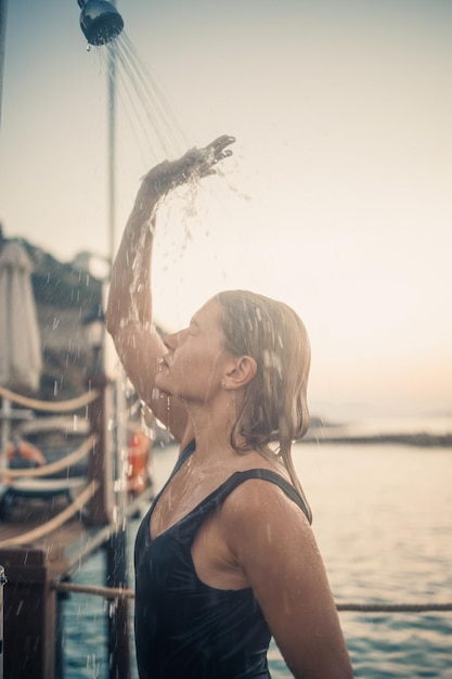 Een mooie jonge aantrekkelijke vrouw in een zwart zwempak met een mooi figuur staat onder een zomerse douche op de pier bij de zee Ze geniet van haar vakantie Selectieve aandacht