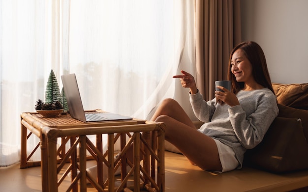 Een mooie jonge aziatische vrouw die een laptopcomputer gebruikt voor videogesprekken terwijl ze thuis koffie drinkt